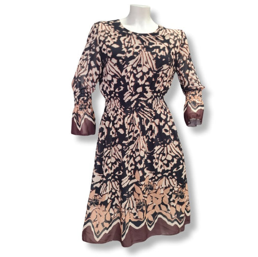 Ein Kleid mit floralen Druckmuster in der Farbe Schoko, Beige, Braun. Rundhals und lange Ärmel. Mit Bordürendruck um Saum und Ämel. 