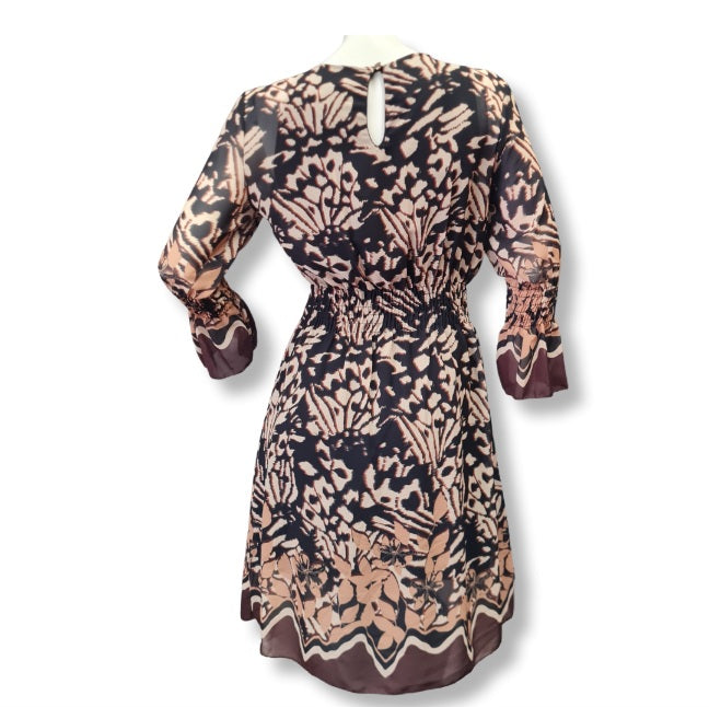 Ein Kleid mit floralen Druckmuster in der Farbe Schoko, Beige, Braun. Rundhals und lange Ärmel. Mit Bordürendruck um Saum und Ämel.  Hier sehen sie die Rückenansicht mit Verschlussknopf
